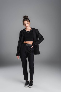 full length of brunette woman in black suit and sneakers standing with hand on hip on grey background