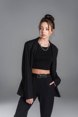 young and stylish woman in black clothing holding hand in pocket and looking away isolated on grey