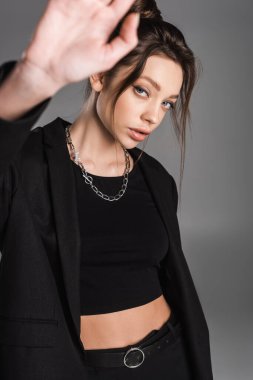 young woman with makeup looking at camera while gesturing with blurred hand isolated on grey