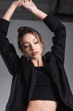 brunette woman in black jacket posing with arms up on grey background