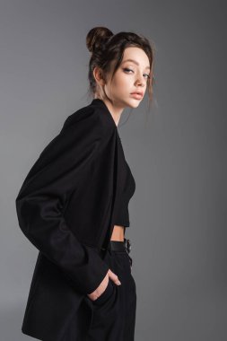 brunette woman in black blazer posing with hand in pocket isolated on grey