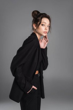 young woman in black clothes standing with hand in pocket and touching face isolated on grey
