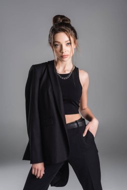 trendy woman in crop top standing with hand in pocket of black trousers isolated on grey