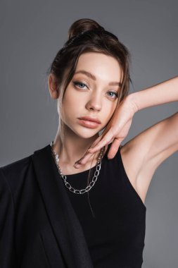 young woman with makeup and silver necklace posing with hand near face isolated on grey