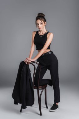 full length of trendy woman in black clothes posing near jacket on chair on grey background