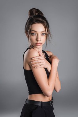 sensual brunette woman looking at camera while embracing herself isolated on grey