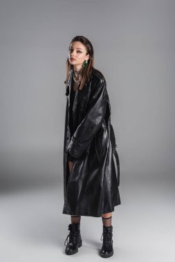 full length of trendy woman in black leather coat on grey background