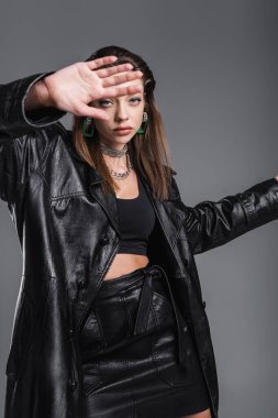 young woman in fashionable leather clothes gesturing near camera on blurred foreground isolated on grey