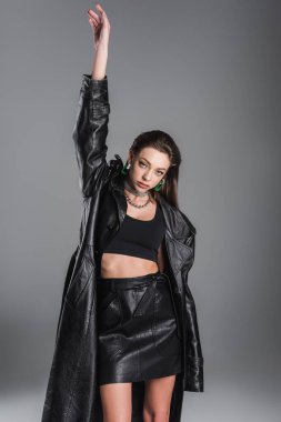 brunette woman in leather coat and black crop top posing with raised hand isolated on grey