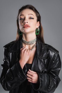 stylish brunette woman with makeup touching silver necklace isolated on grey