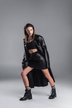full length of woman in black leather skirt and coat looking away while posing on grey background