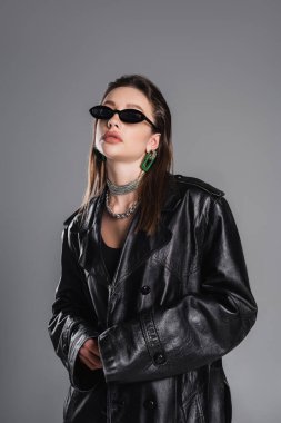 brunette woman in trendy sunglasses and black leather coat posing isolated on grey