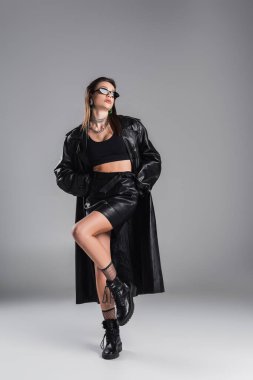 full length of young woman in black leather clothing and dark sunglasses posing on one leg on grey background