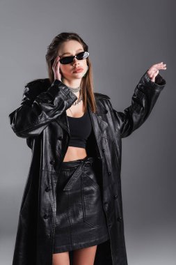 young brunette woman in black leather coat touching stylish sunglasses isolated on grey