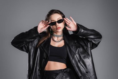 young woman in silver necklaces and black clothing adjusting dark sunglasses isolated on grey