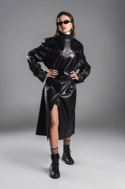 full length of woman wearing black leather coat back to front and posing with hands on hips on grey background