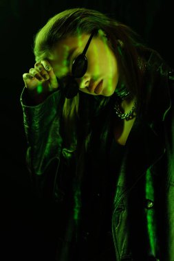 young woman in sunglasses and leather coat posing with bowed head isolated on black