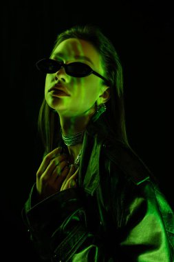 portrait of brunette woman in trendy sunglasses and silver necklaces in green light isolated on black
