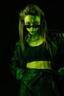 trendy woman in crop top looking at camera over dark sunglasses in green light isolated on black clipart