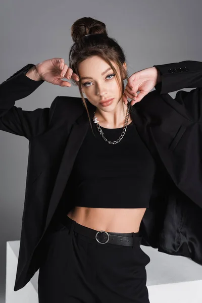 sensual woman in black crop top and blazer posing with hands near face isolated on grey