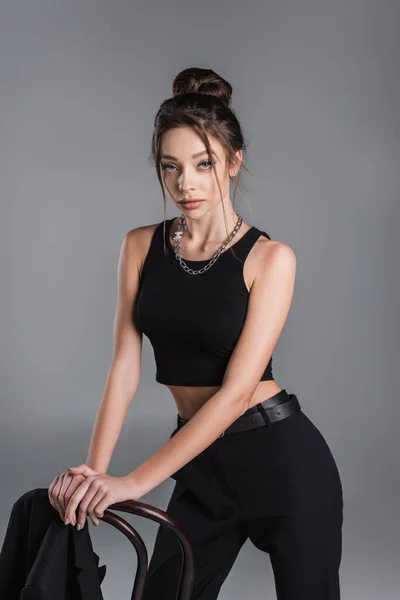 trendy woman in black crop top and silver necklace posing near chair isolated on grey