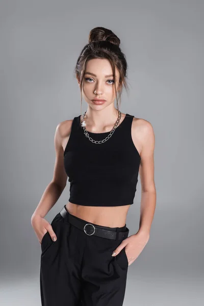 young woman in black crop top posing with hands in pockets of trousers isolated on grey