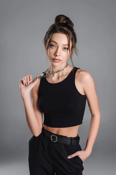 fashionable woman in black crop top touching metal necklace and looking at camera isolated on grey