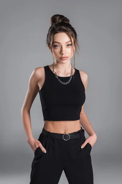 brunette woman in black crop top posing with hands in pockets isolated on grey
