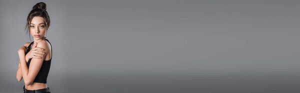 young woman in black crop top hugging herself and looking at camera isolated on grey, banner