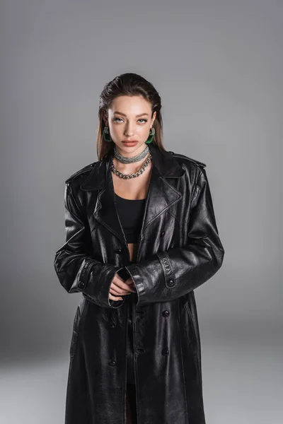 brunette woman in black leather coat and silver accessories looking at camera isolated on grey