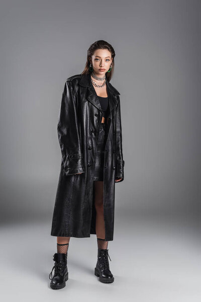 full length of young woman in leather boots and black coat looking at camera on grey background