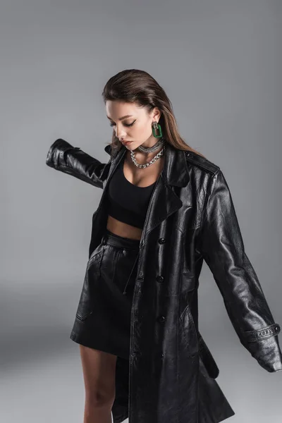 trendy woman in black leather coat and silver accessories posing isolated on grey