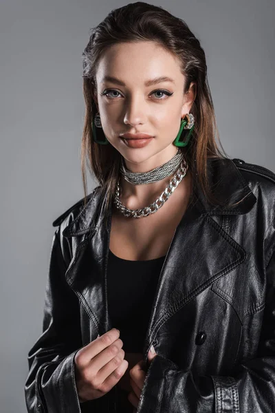 positive and trendy woman with makeup and silver accessories looking at camera isolated on grey