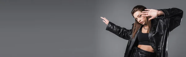 pretty woman in black crop top and leather coat gesturing and looking at camera isolated on grey, banner