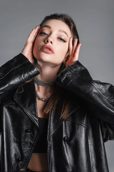 brunette woman in black leather coat and makeup touching face isolated on grey