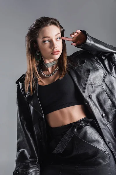 brunette woman in crop top and black leather coat looking away isolated on grey