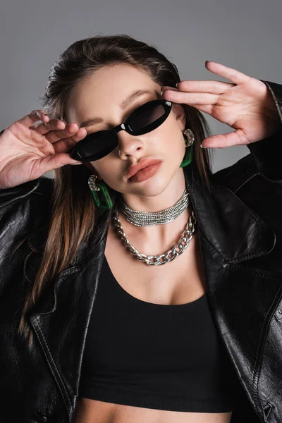 young woman in silver necklaces adjusting dark and trendy sunglasses isolated on grey
