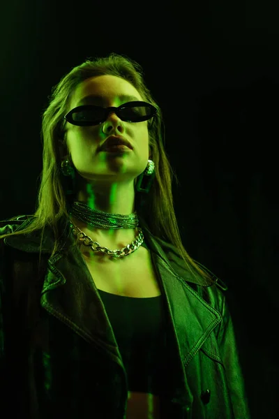 low angle view of young woman in trendy sunglasses and metal necklaces in green neon light isolated on black
