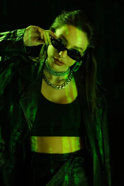 trendy woman in crop top holding sunglasses and looking at camera in green light isolated on black