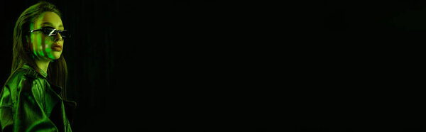 portrait of brunette woman in dark sunglasses and leather coat in green light isolated on black, banner