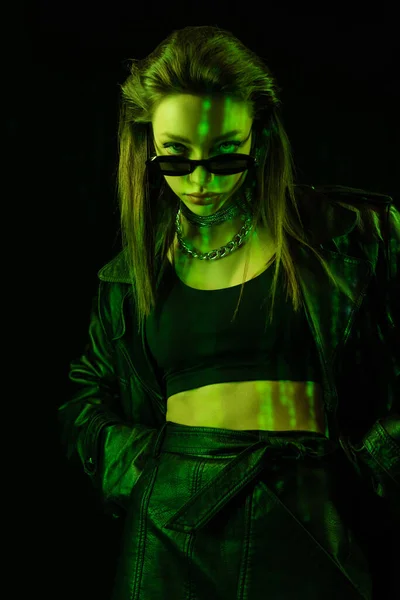 trendy woman in crop top looking at camera over dark sunglasses in green light isolated on black