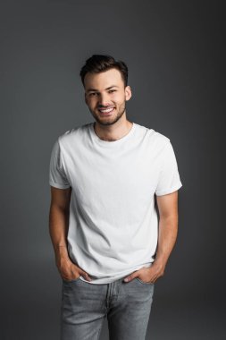Positive brunette man in white t-shirt and jeans posing isolated on grey 