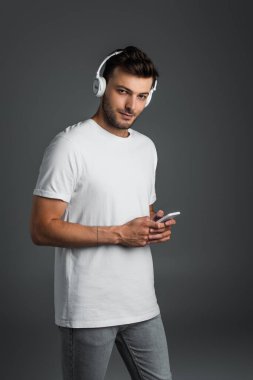 Young man using wireless headphones and smartphone and looking at camera isolated on grey 
