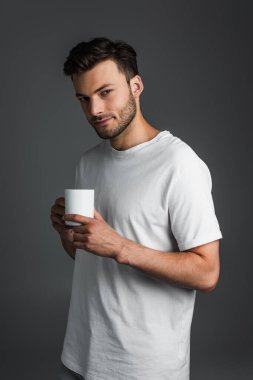 Brunette man in white t-shirt holding cup and looking at camera isolated on grey 