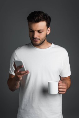 Young man holding cup of coffee and using smartphone isolated on grey 