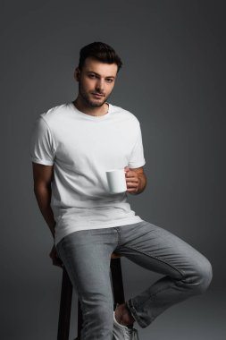 Young man in white t-shirt and jeans holding coffee cup while sitting on chair isolated on grey 