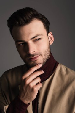 Brunette man in jacket touching chin and looking away isolated on grey 