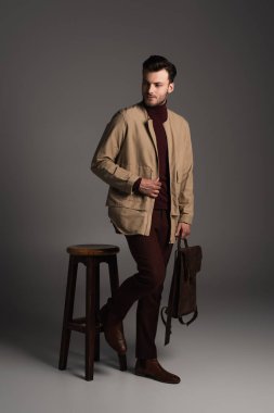 Stylish man in brown autumn jacket holding backpack near wooden chair on grey background