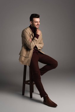 Full length of smiling man in autumn outfit sitting on chair on grey background