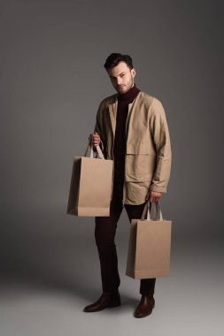 Full length of trendy man in autumn jacket holding purchases on grey background